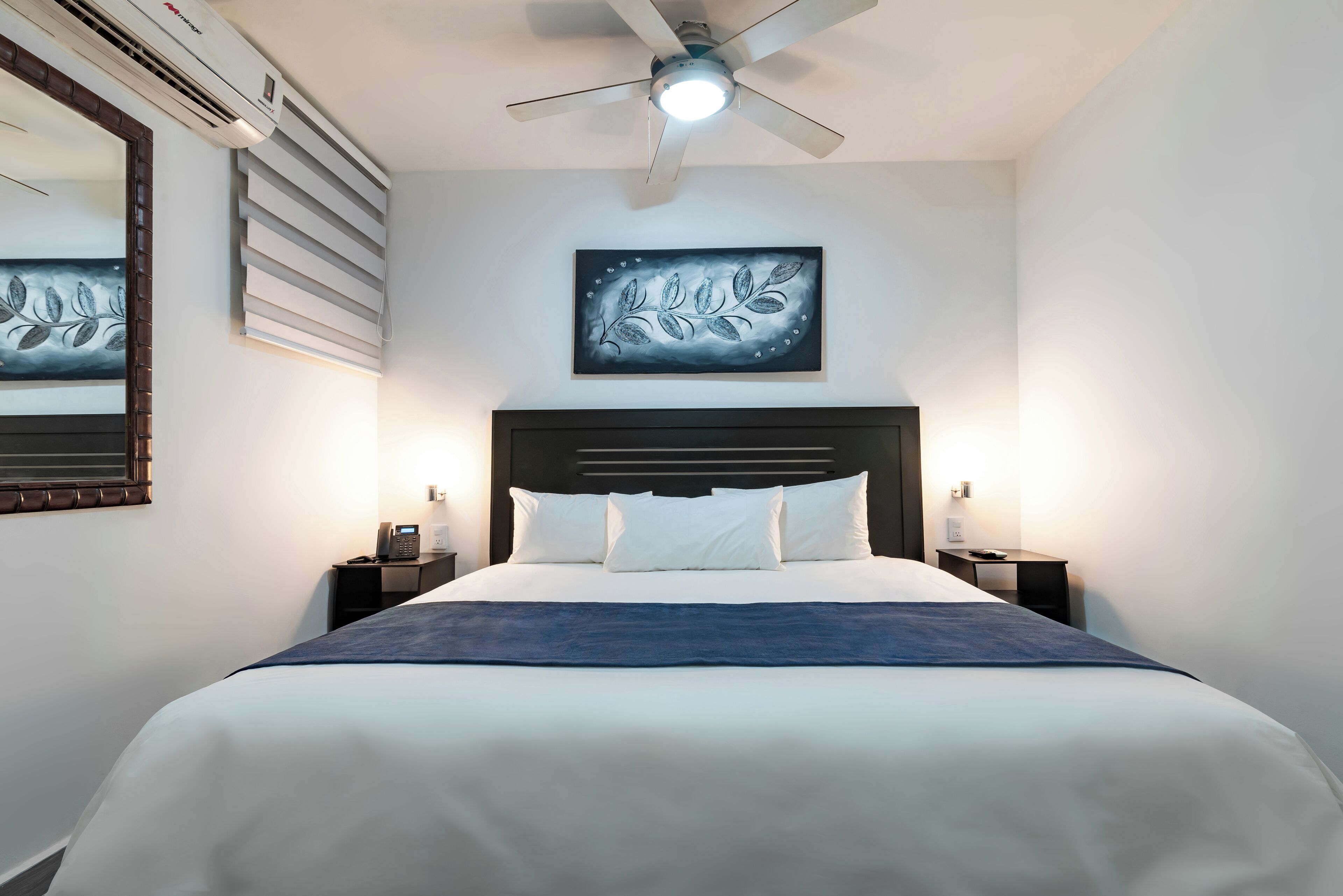 A hotel room with a bed, a ceiling fan, an air conditioning unit, a bedside table with a phone, and a wall-mounted artwork.