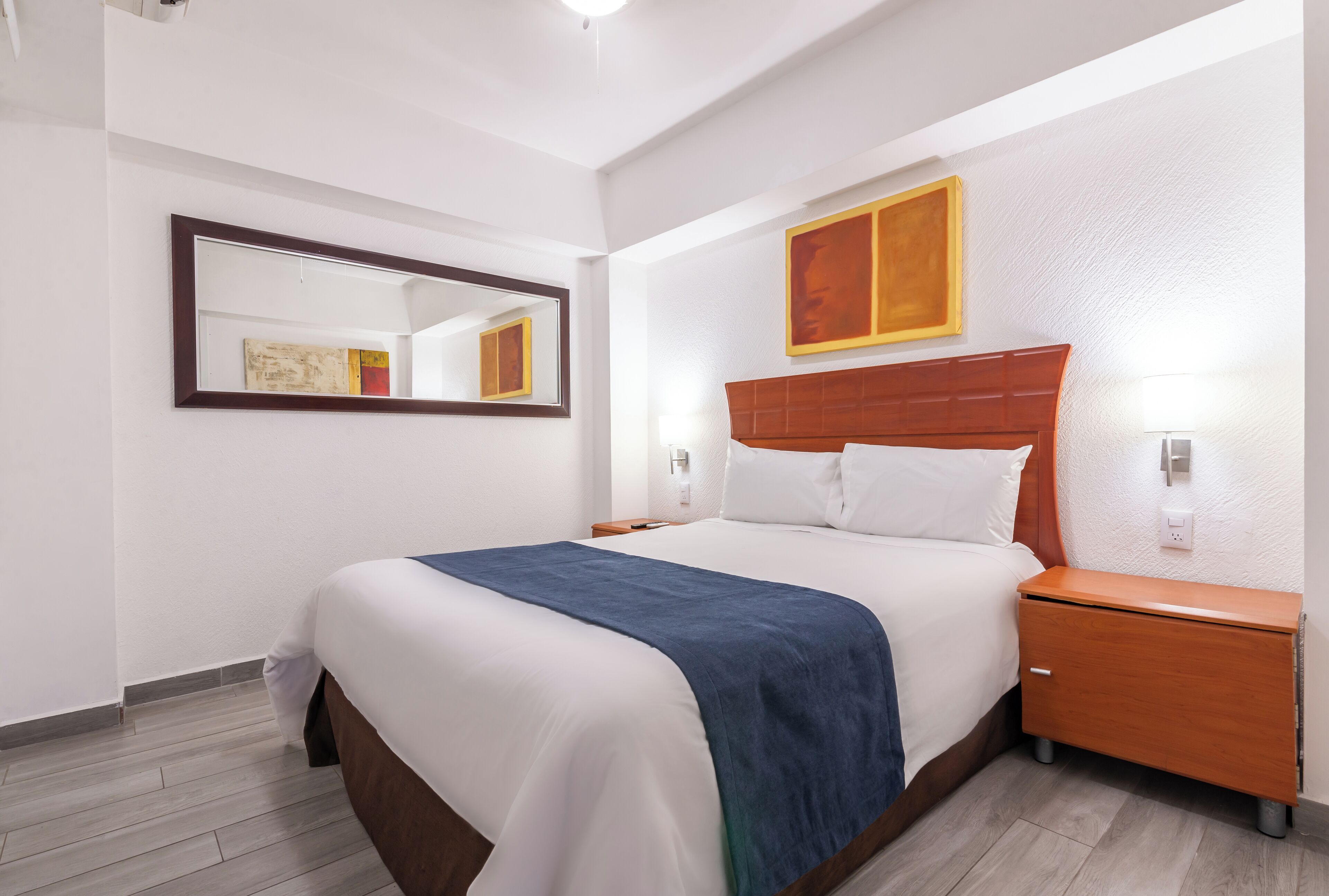 A hotel room with a large bed, a wooden nightstand, a mirror, and a painting on the wall.