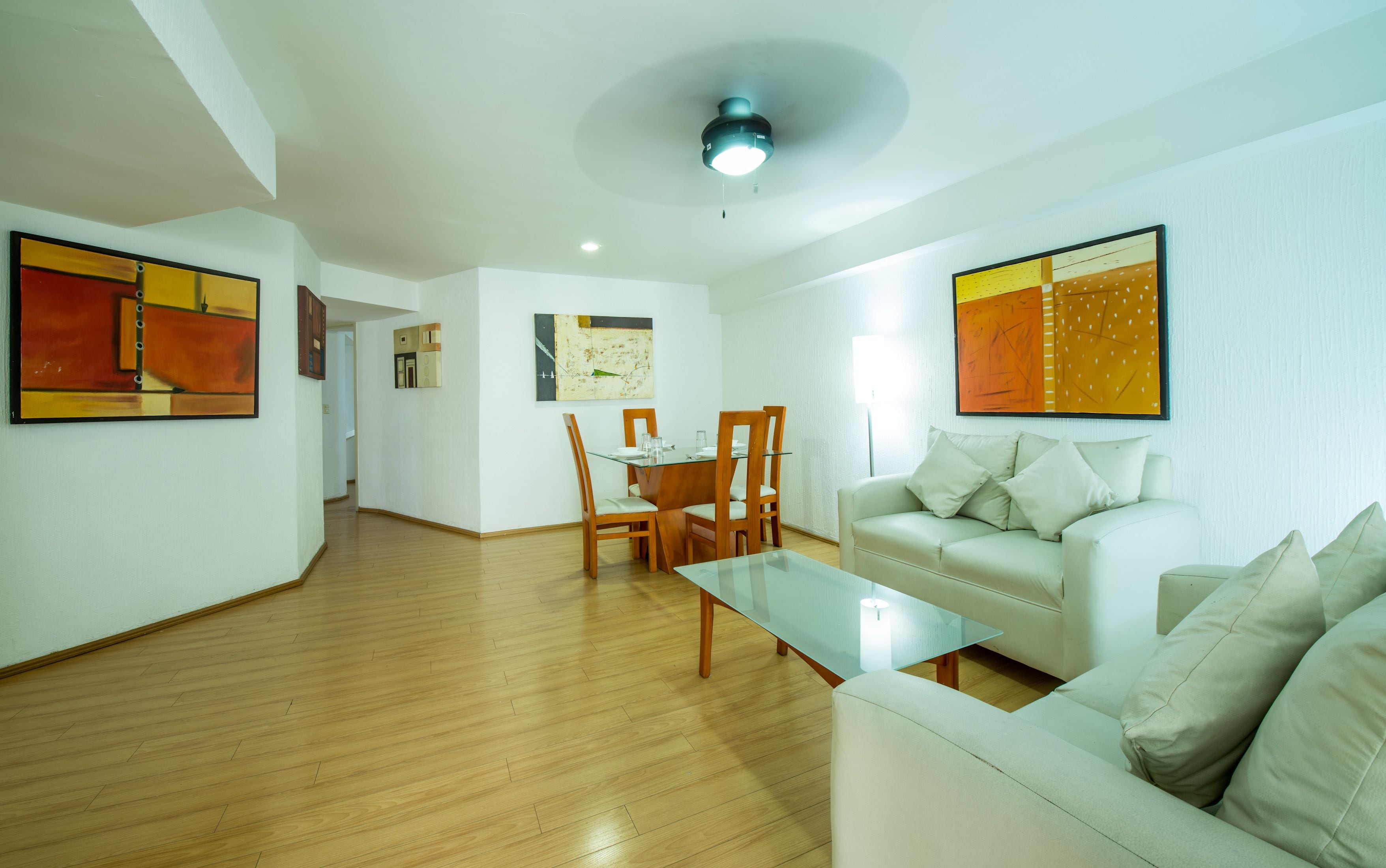 A spacious living room with a wooden floor, a glass coffee table, a white sofa, and a dining area with wooden chairs and a glass table.