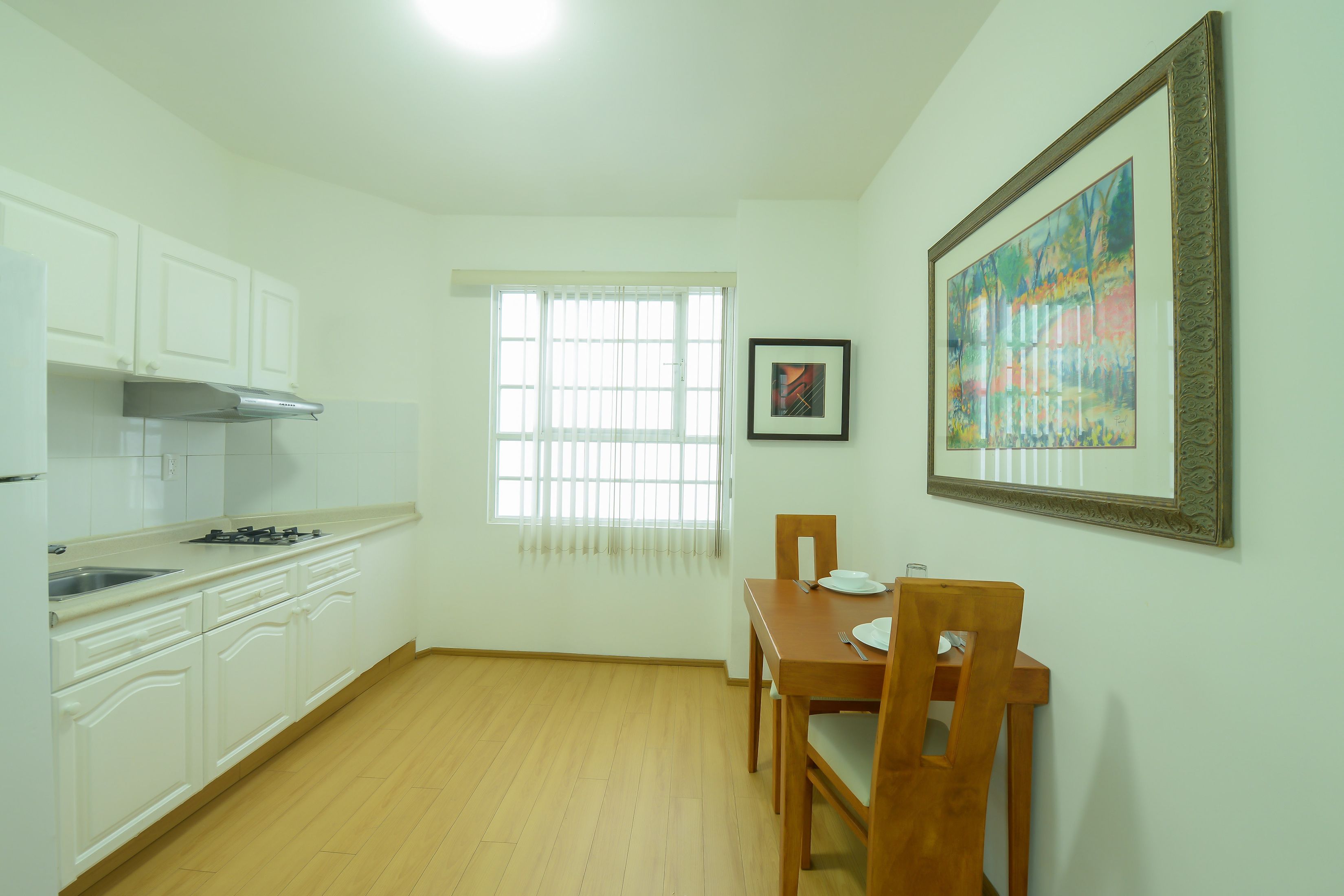 A kitchen with white cabinets, a dining table with chairs, a window with blinds, and a framed painting on the wall.