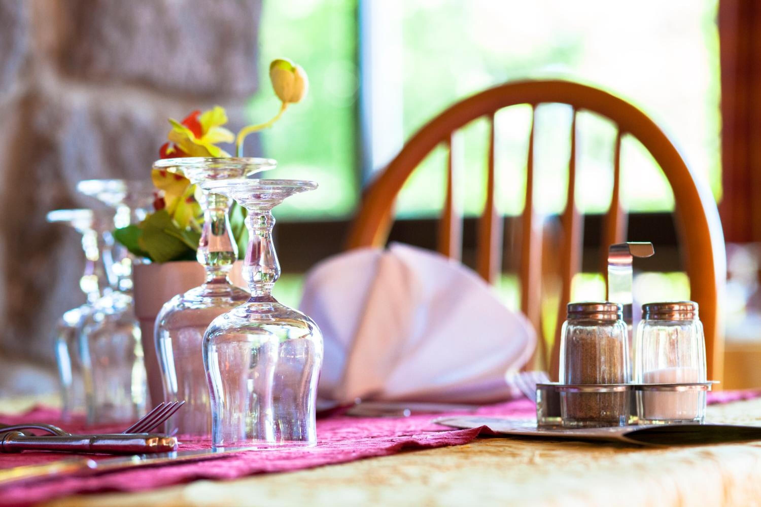 Uma mesa de jantar posta com copos de vidro transparente, saleiro e pimenteiro, e uma flor em um vaso de vidro.
