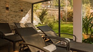 A modern lounge area with wicker chairs and a stone wall, overlooking a garden and a bridge. - Hotel Adler (Nova Levante)