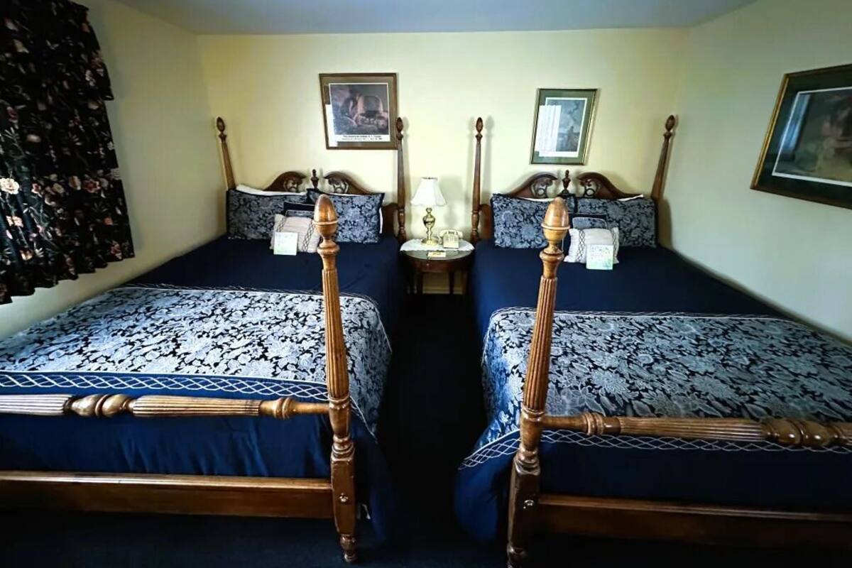 Quarto com duas camas, cada uma com estrutura de madeira e roupa de cama estampada. Há mesas de cabeceira com abajures e quadros nas paredes.