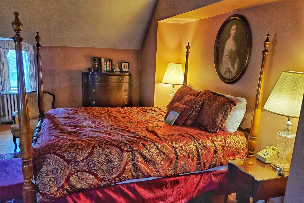 Quarto com cama de dossel, cômoda, escrivaninha com abajur e retrato emoldurado na parede.