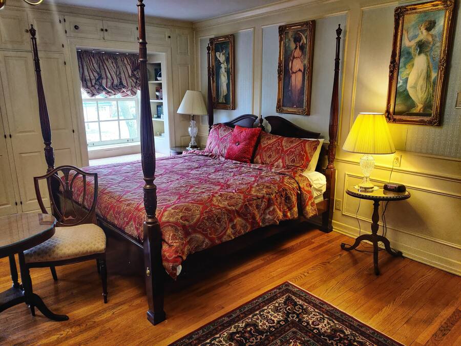 A four-poster bed with a patterned bedspread, a chair, a bedside table with a lamp, and a rug on a wooden floor.