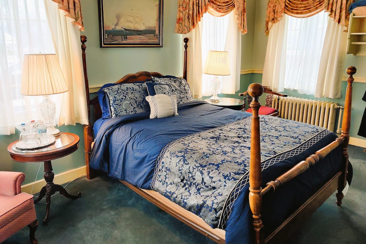 Uma cama de dossel clássica com colcha floral, uma mesa de cabeceira redonda de madeira com uma luminária e uma pintura emoldurada com tema marítimo na parede.