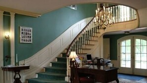 A grand staircase with a chandelier, a desk with a chair, and a large rug in a spacious room with teal walls. - Montague Inn (Saginaw)