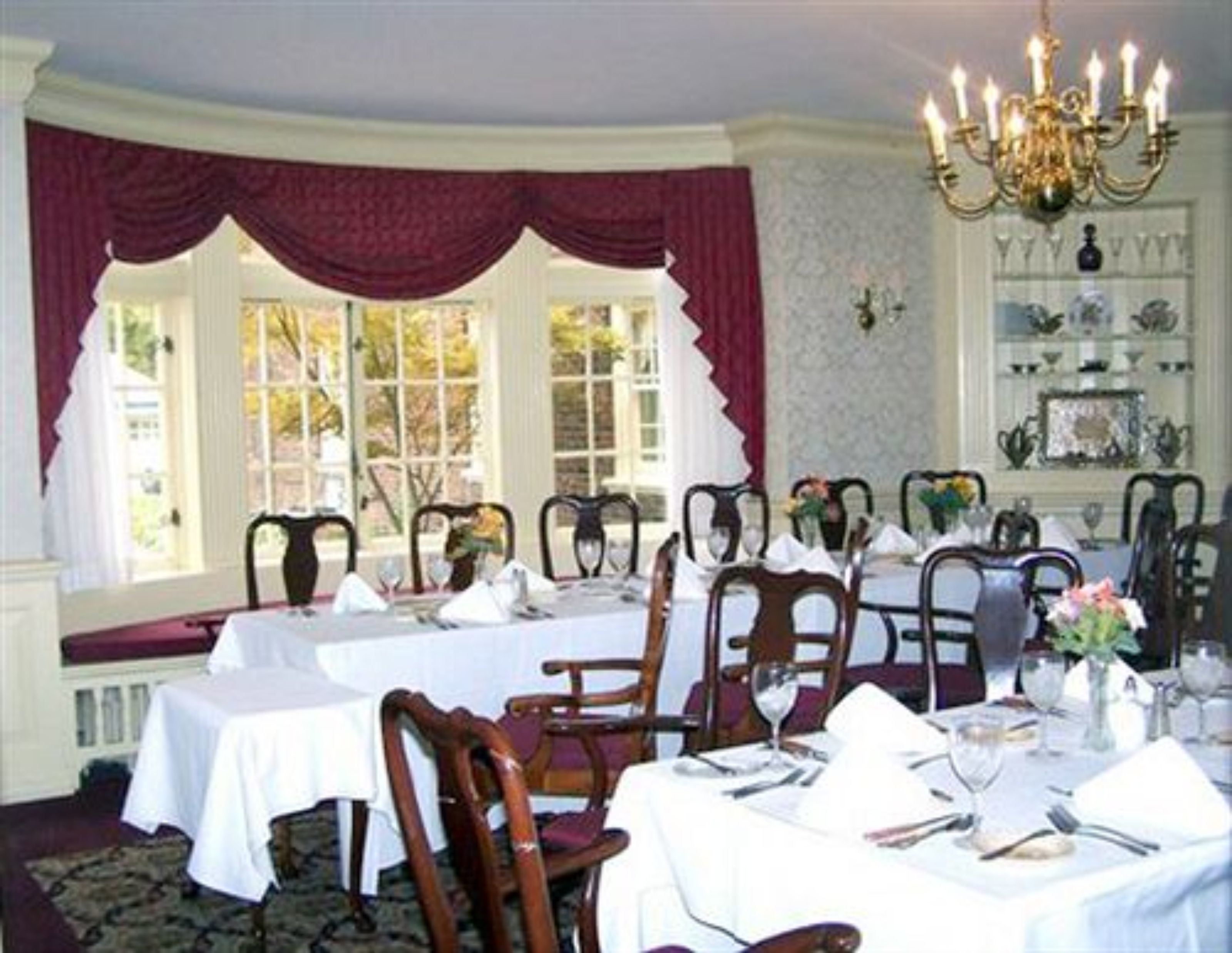 A dining room with tables set for a meal, chairs, a chandelier, and a view of the outdoors through large windows.