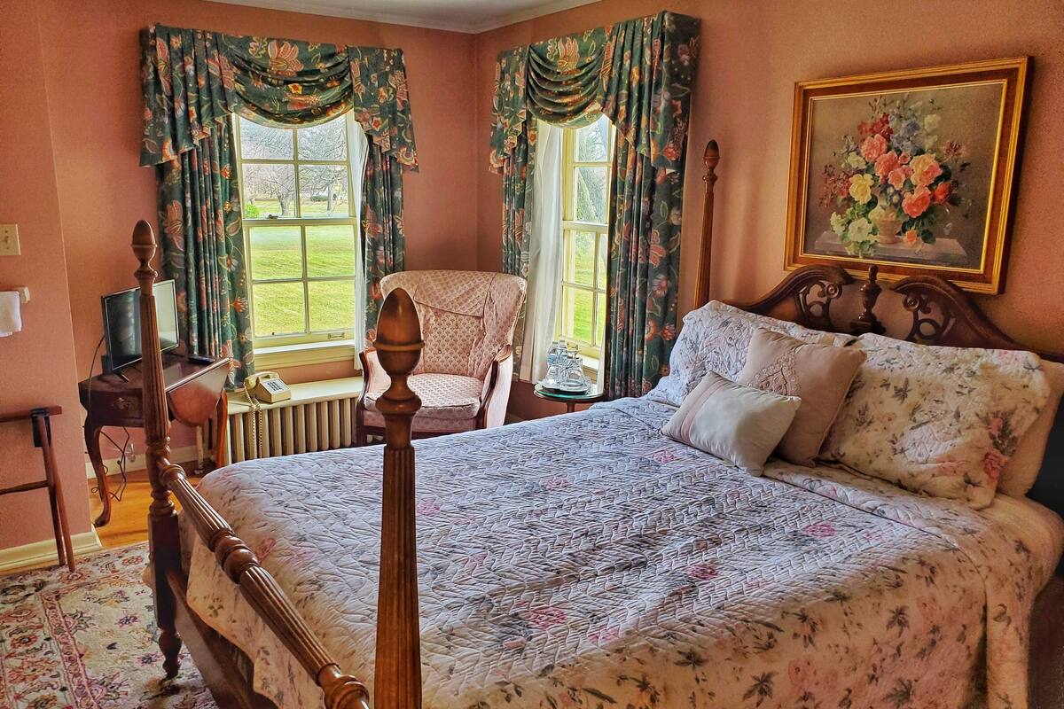 Quarto com cama de dossel, uma cadeira de madeira, escrivaninha com computador e janela com cortinas.