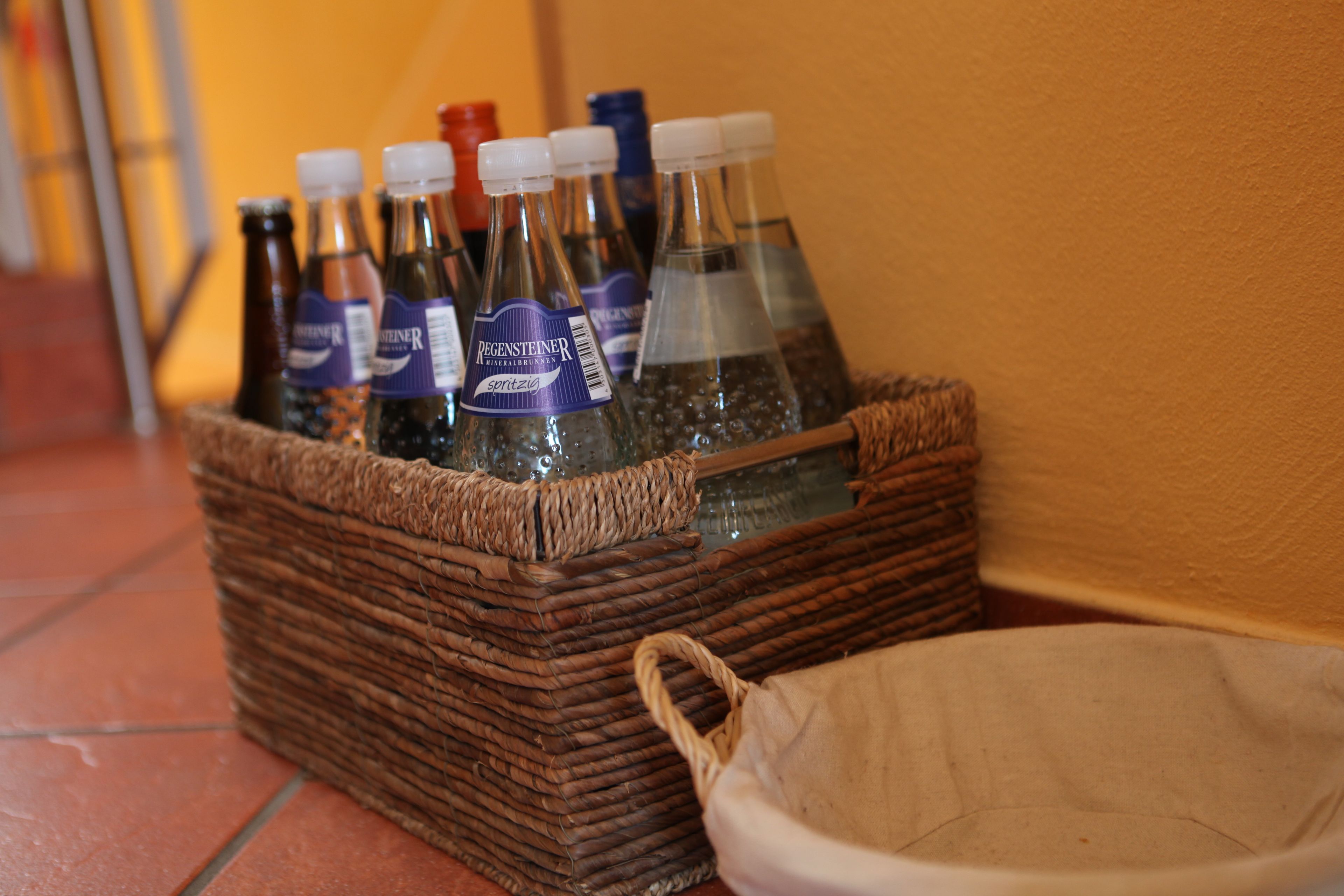 A wicker basket containing several bottles of beverages, a small cloth bag, and a ceramic bowl on a tiled floor.