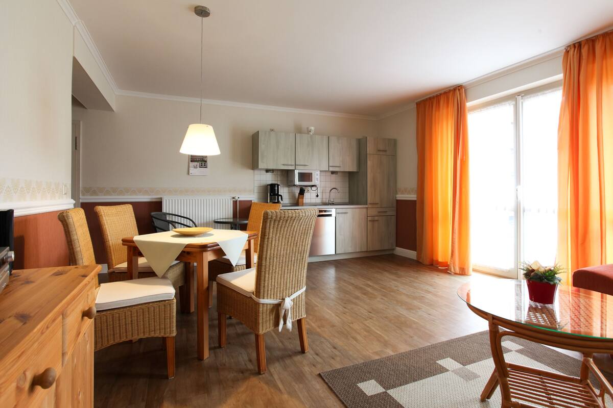 Eine moderne Küche mit hellen Holzschränken, einer Mikrowelle und einer Kaffeemaschine. Es gibt einen Essbereich mit Korbstühlen und einem Tisch, sowie eine kleine Sitzecke mit einem Glastisch und orangefarbenen Vorhängen.