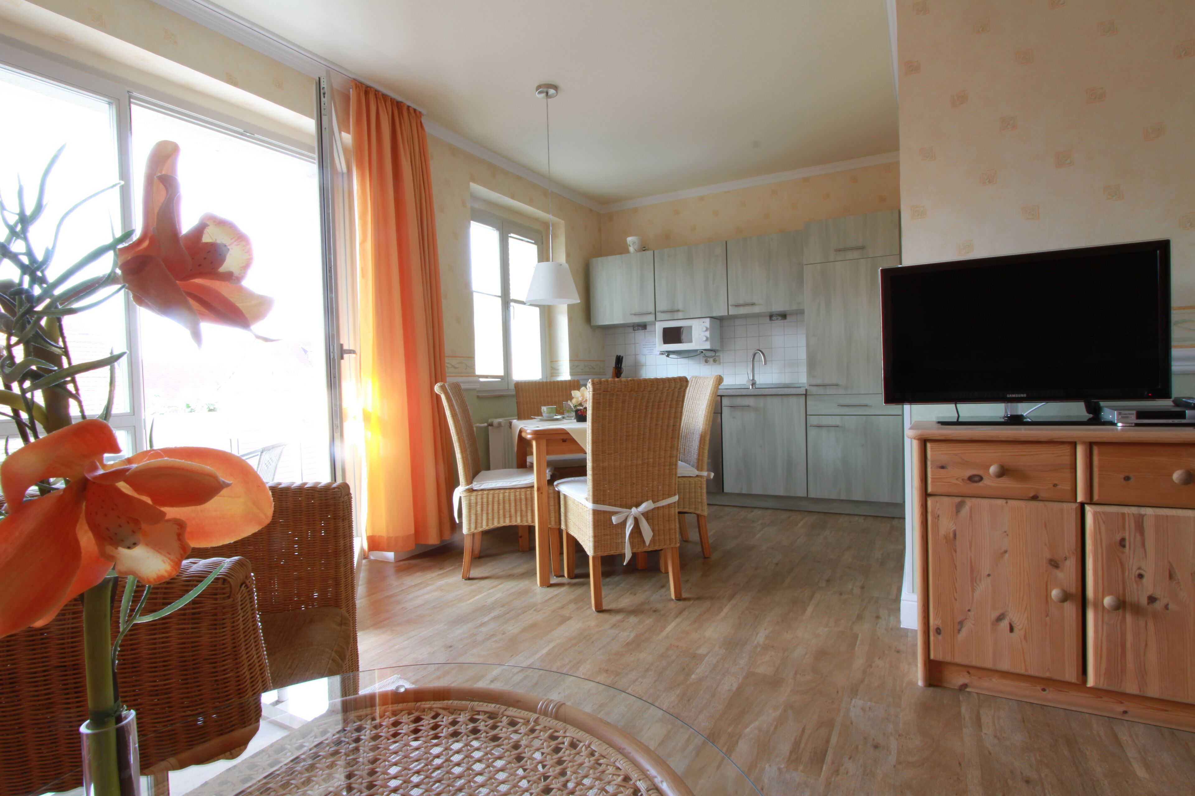 A dining area with wicker chairs and a table, a kitchen with light-colored cabinets, and a television on a wooden cabinet.