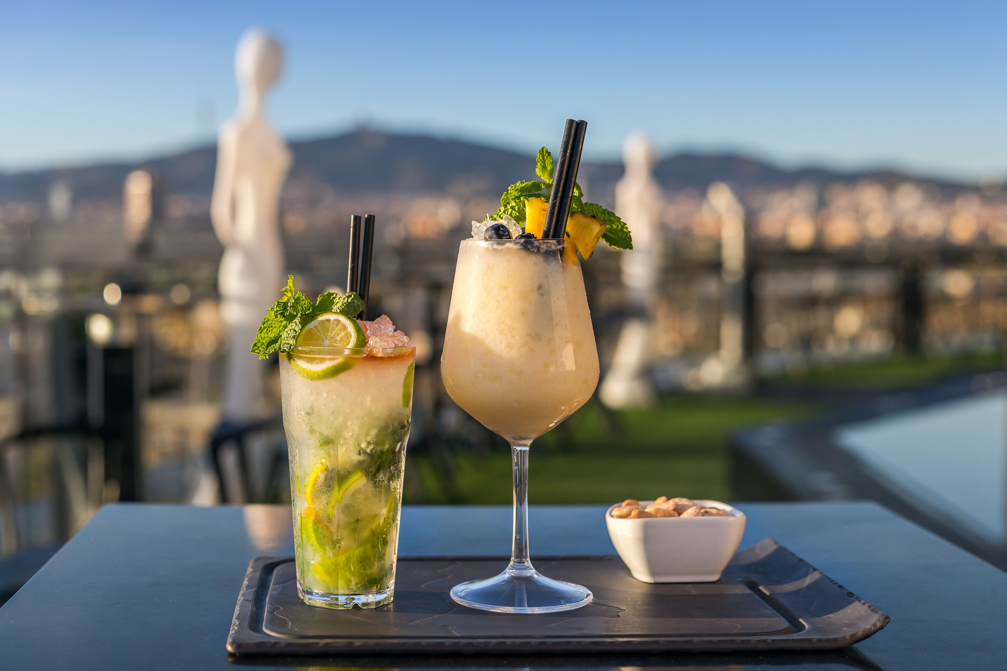 Two drinks on a table with a cityscape and statues in the background.