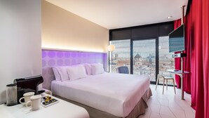 A hotel room with a large bed, a coffee machine, a table with a chair, and a view through a window with a metal fence.