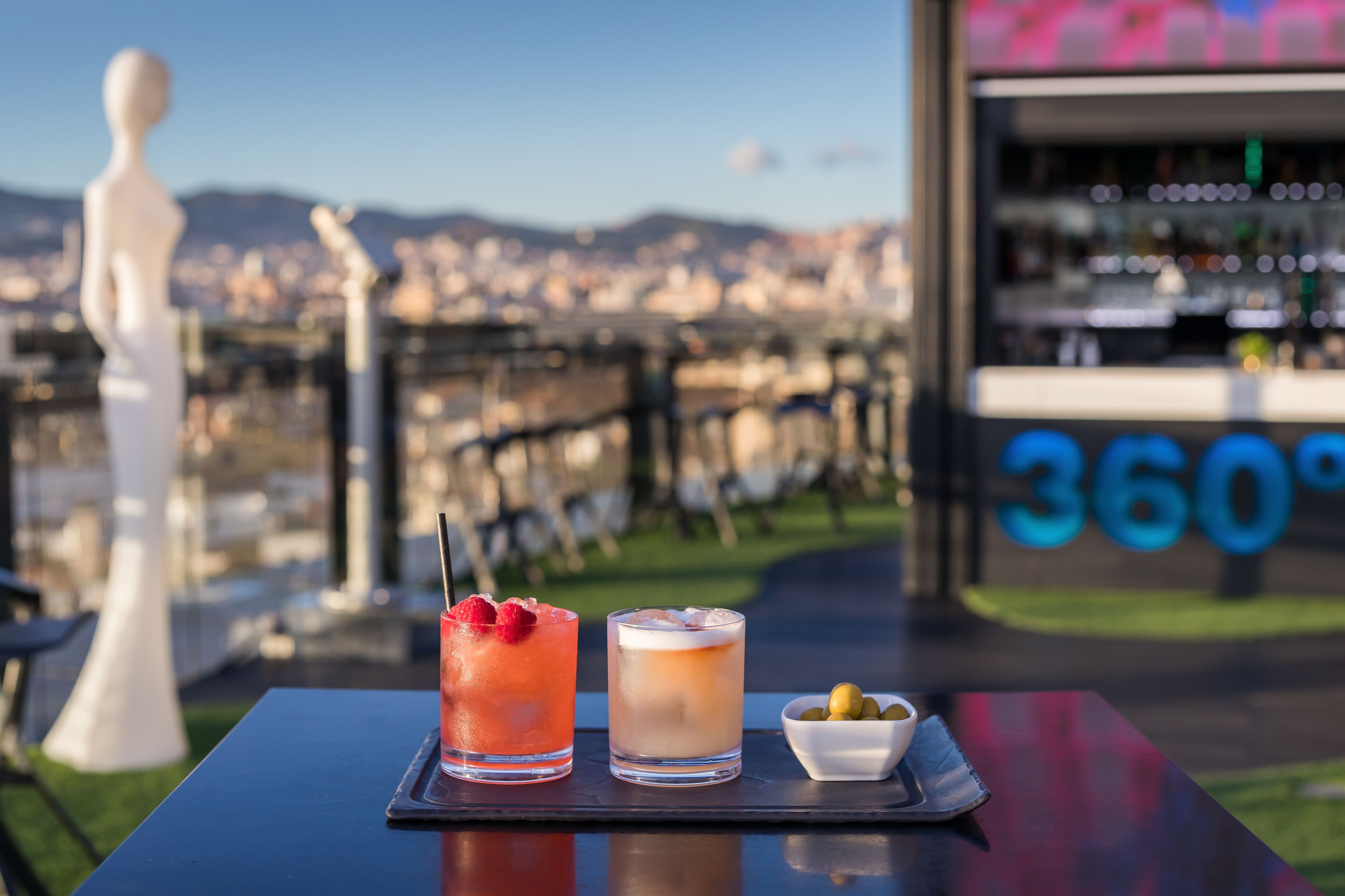 A table with two drinks and a small bowl of olives, set against a cityscape view.