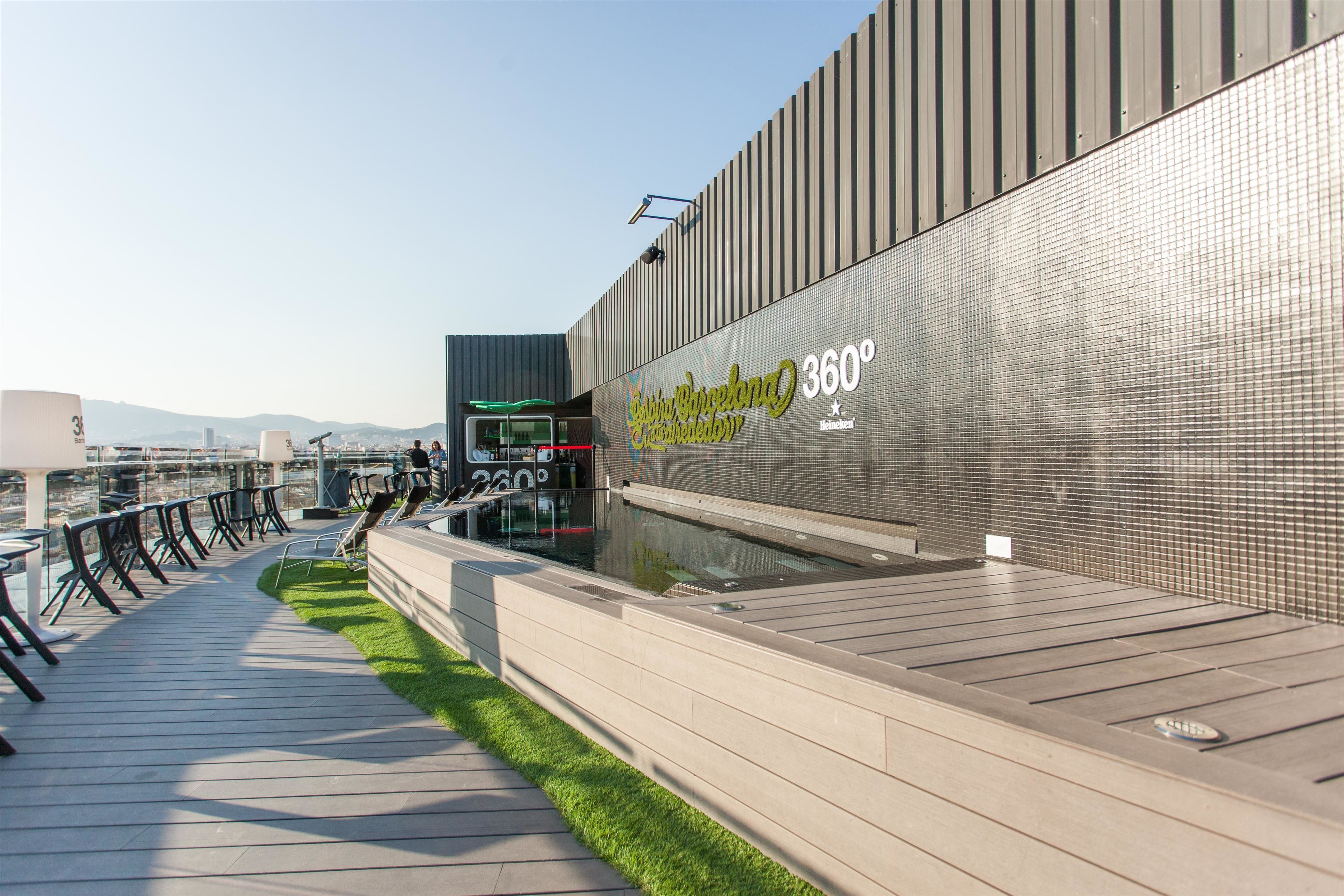 A modern rooftop terrace with a glass balustrade, a water feature, and a seating area with chairs and tables.