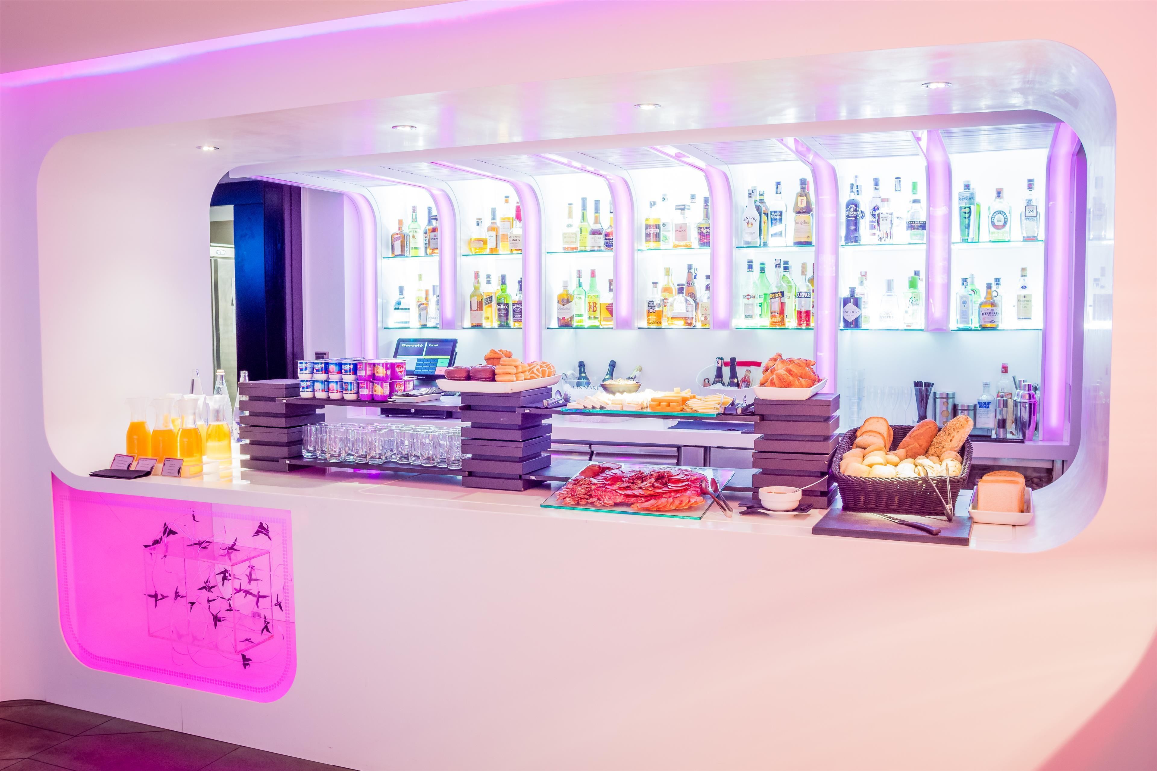 A bar area with shelves of beverages, a counter with snacks, and a decorative wall with a pink light.