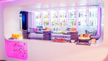 A bar area with shelves of beverages, a counter with snacks, and a decorative wall with a pink light.