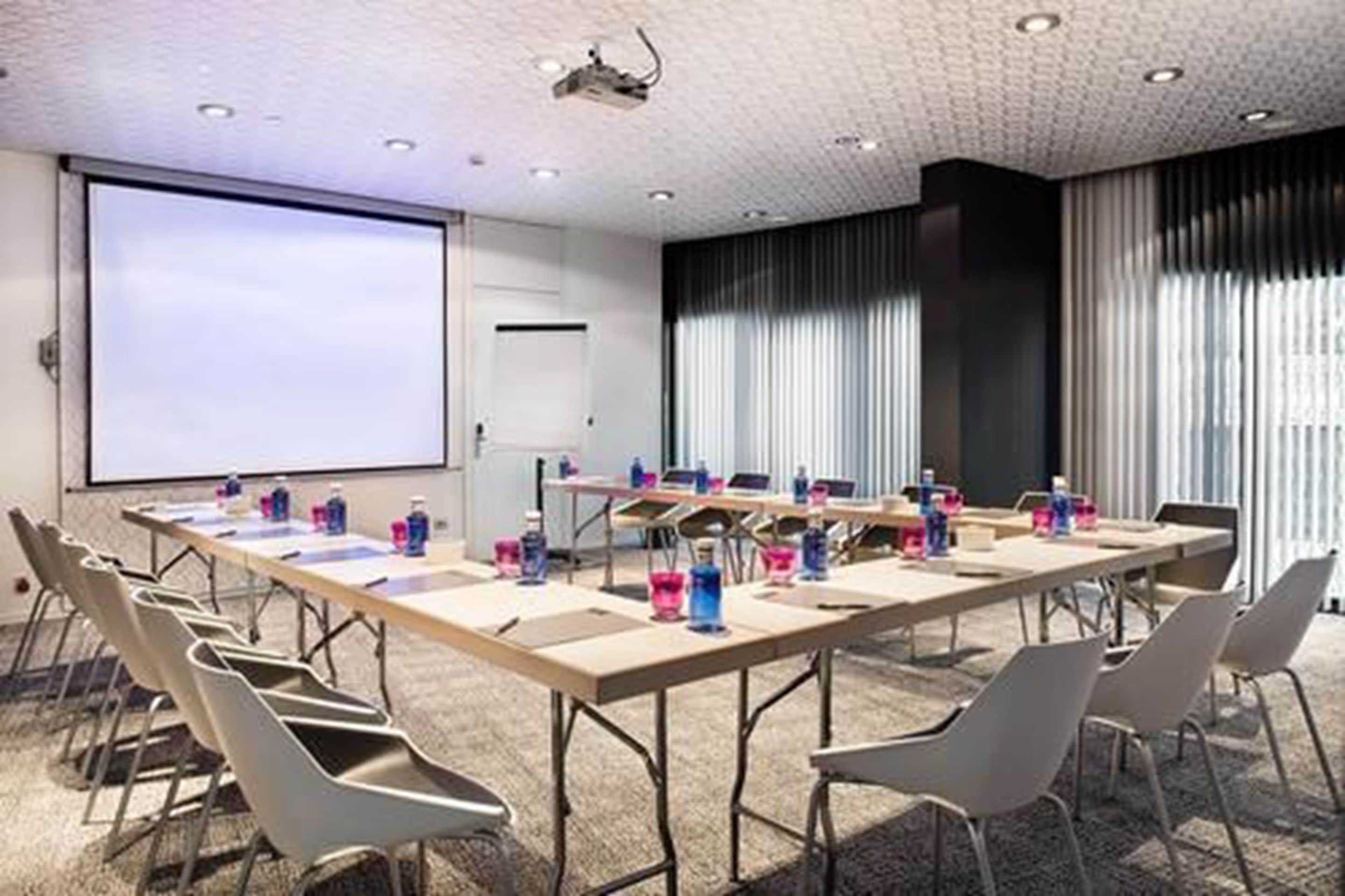 Een congreszaal met een lange tafel, stoelen, een projectiescherm en een groot raam met gordijnen.