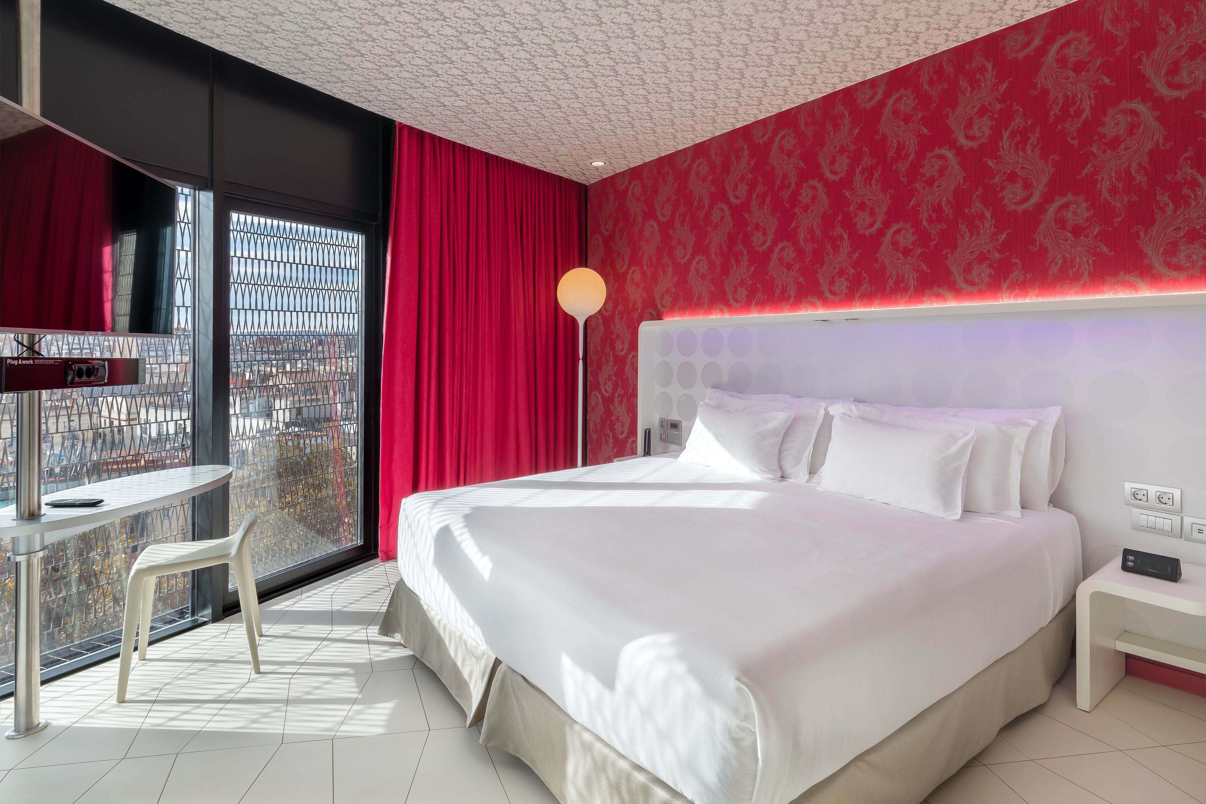 A hotel room with a large bed, a desk with a chair, a standing lamp, and a view through a window with red curtains.
