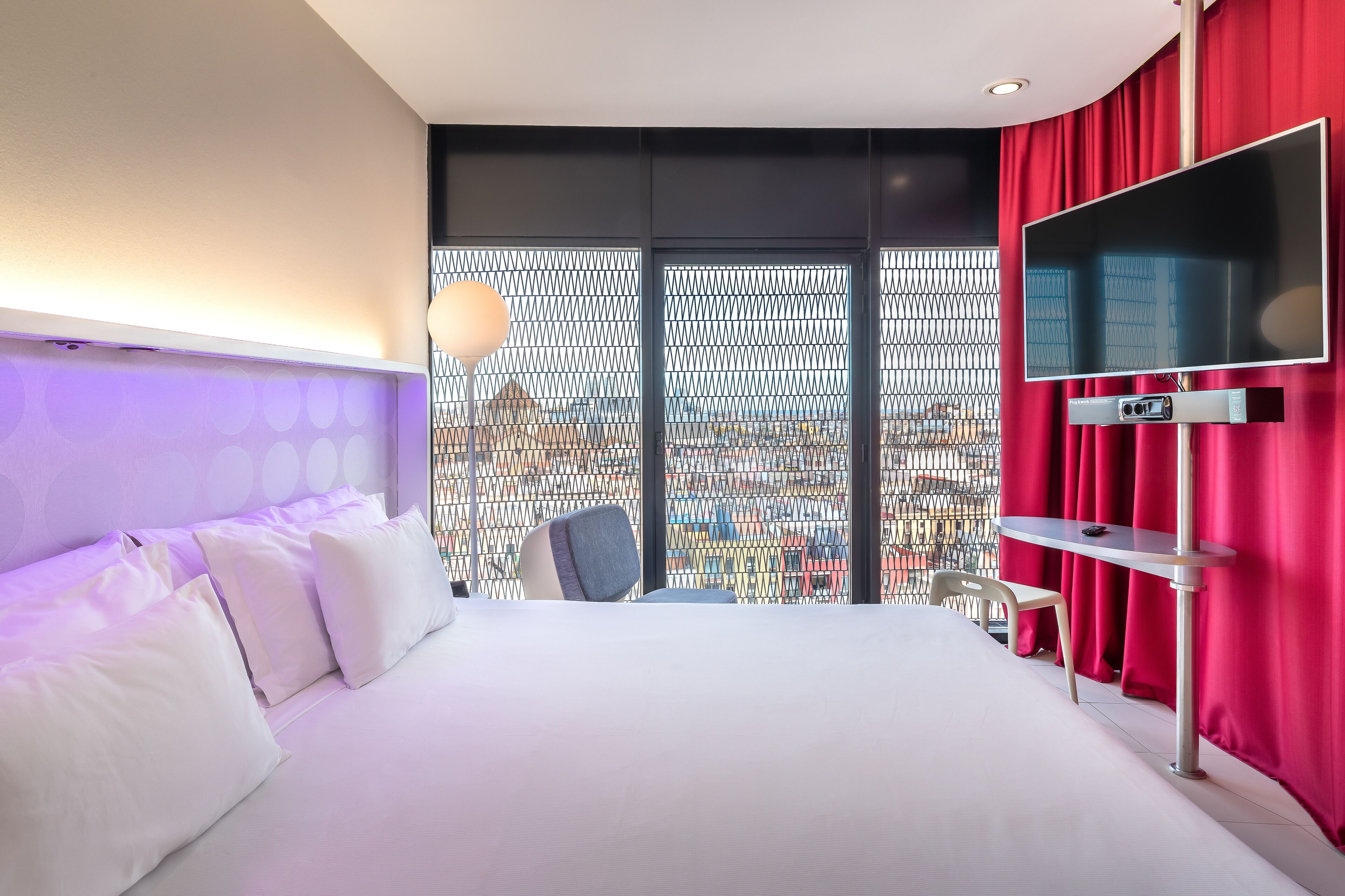 A hotel room with a large bed, a TV mounted on the wall, a desk with a chair, and a view of the city through a window with a metal mesh screen.