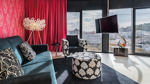 A modern living room with a dark blue sofa, patterned cushions, a round ottoman, a floor lamp, and a flat-screen TV. - Barceló Raval (Barcelona)