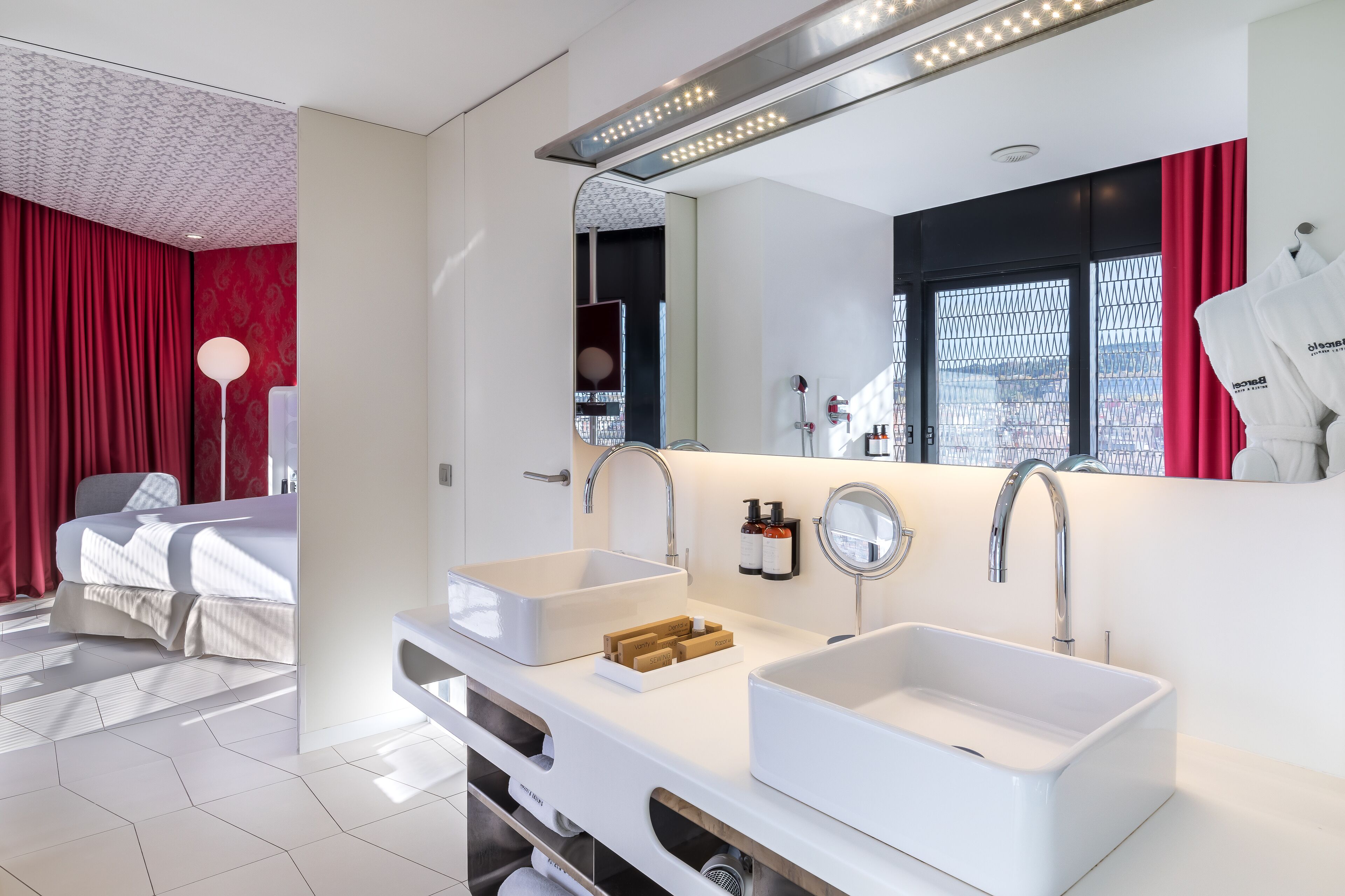 A modern hotel room with a large bed, a white vanity with two sinks, a mirror, and a window with red curtains.