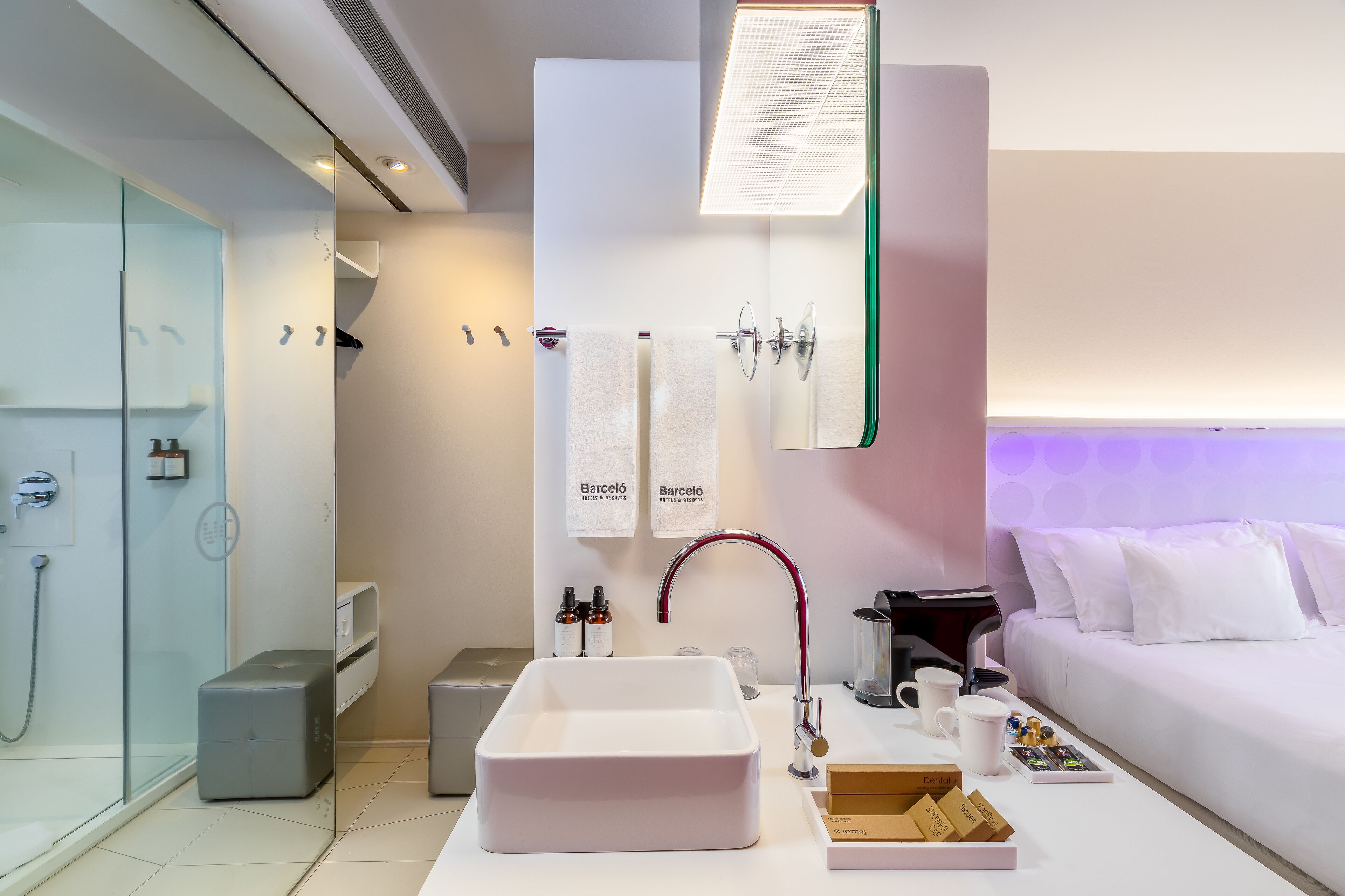 A modern bathroom with a large sink, a glass shower enclosure, and a bed with white bedding.