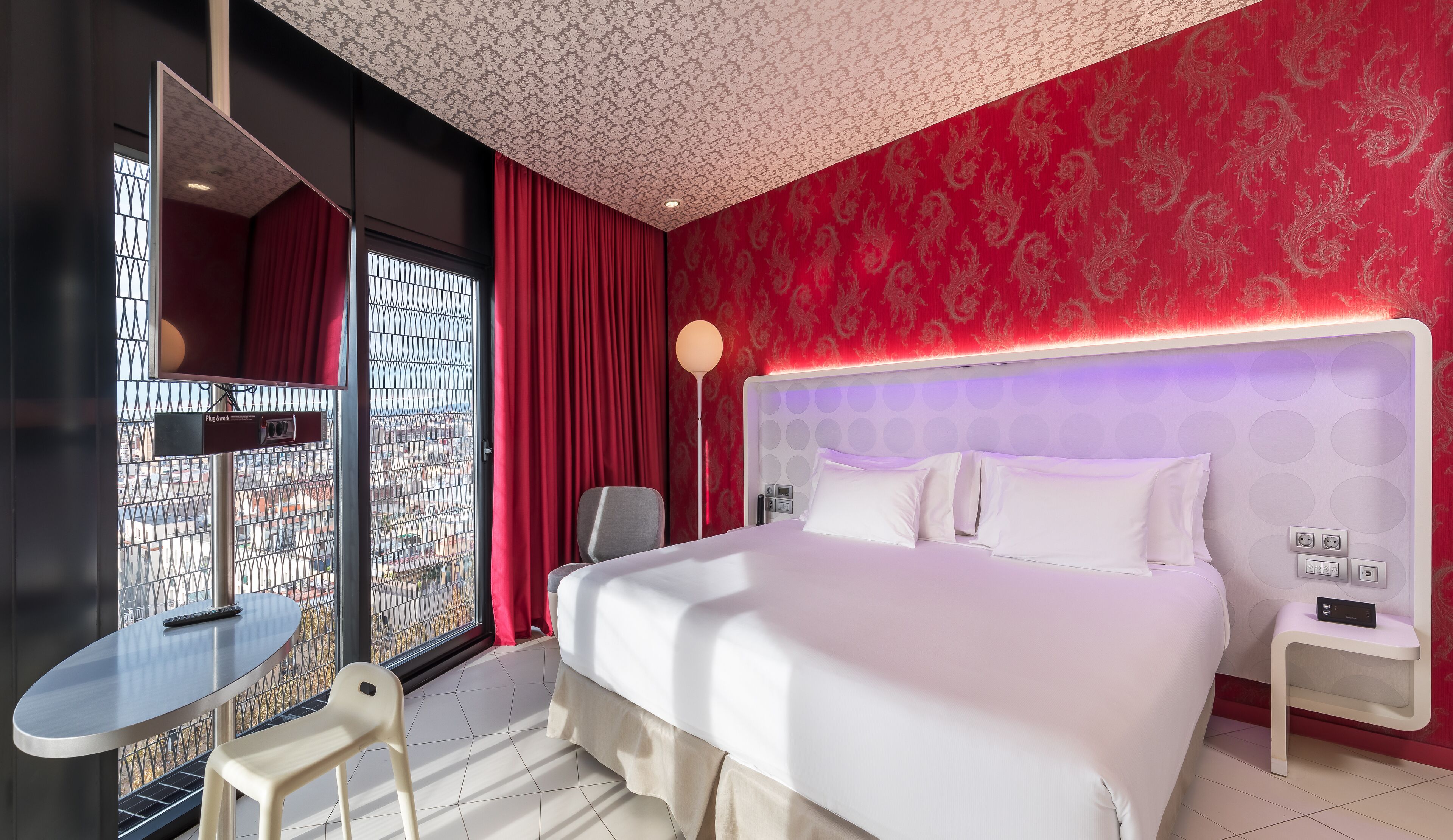 A hotel room with a large bed, a desk, a chair, and a view through a window with metal bars.