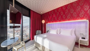 A hotel room with a large bed, a desk, a chair, and a view through a window with metal bars.