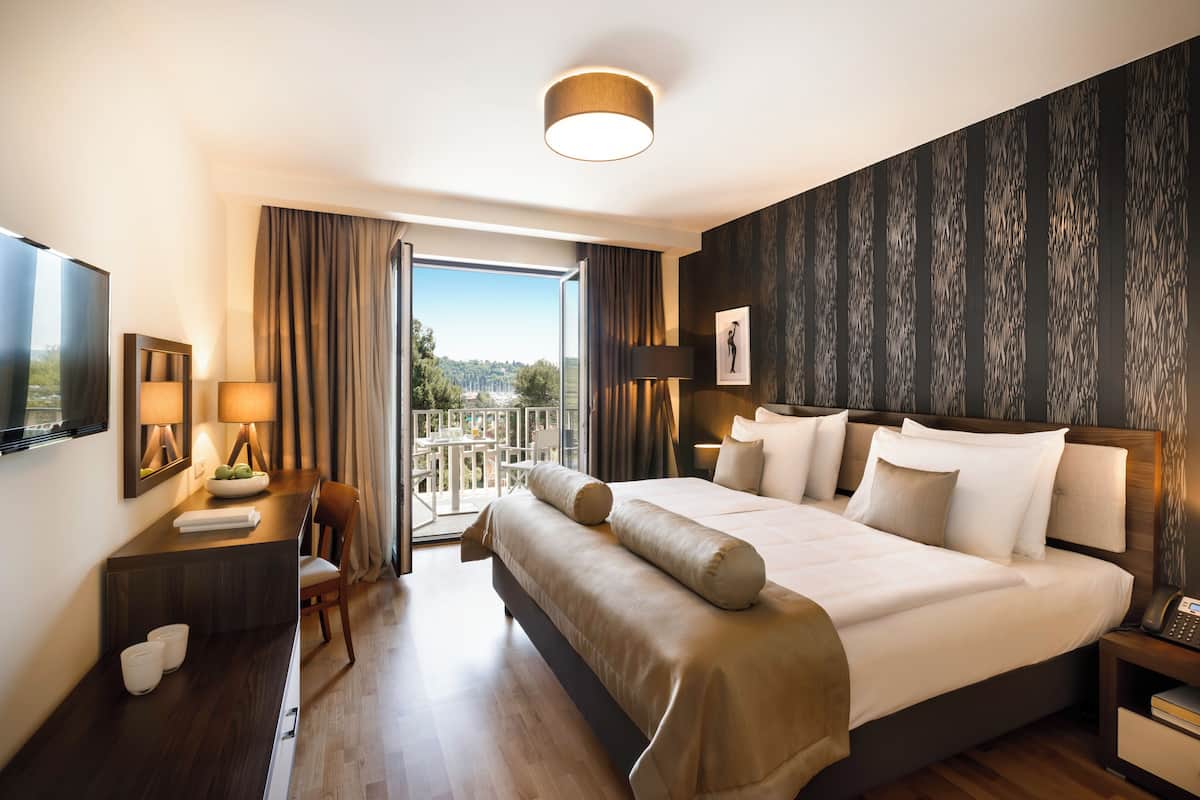 Habitación de hotel con una cama grande, un escritorio con silla, televisor y un balcón con vistas.