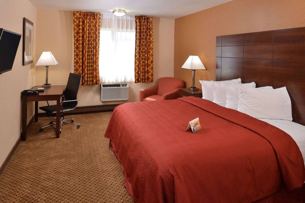 A hotel room with a large bed, two chairs, a desk, a TV, and an air conditioning unit.