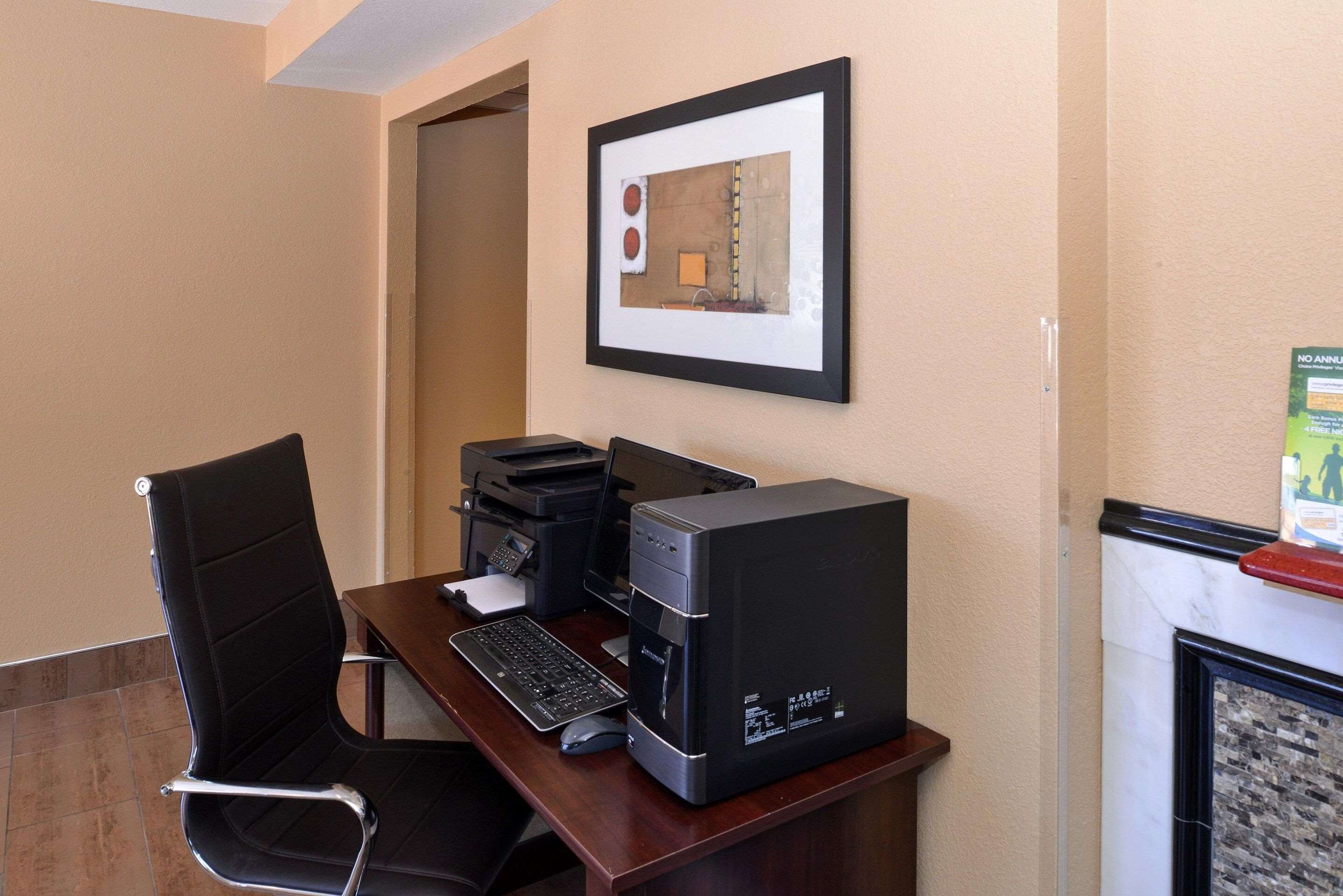 Una habitación de hotel con un escritorio, una silla negra, un ordenador de sobremesa, una impresora y un cuadro en la pared.