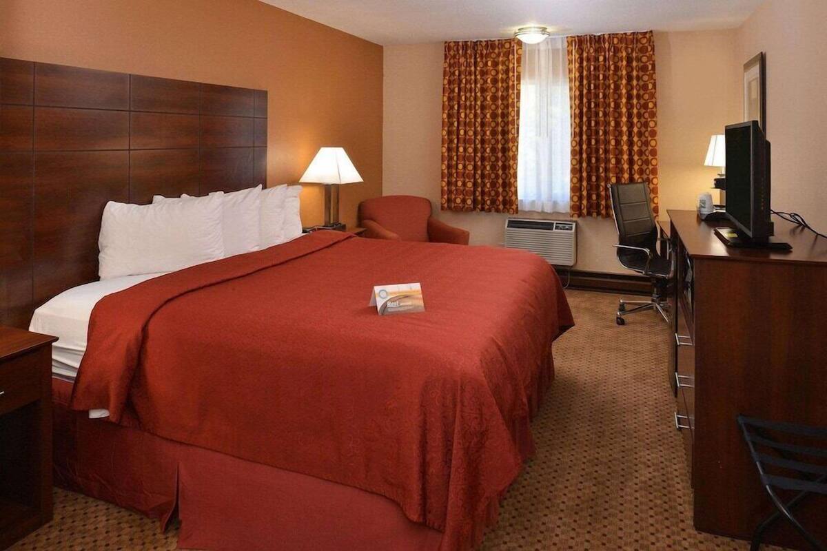 A hotel room with a large bed, a desk with a computer, a chair, a lamp, and a window with curtains.