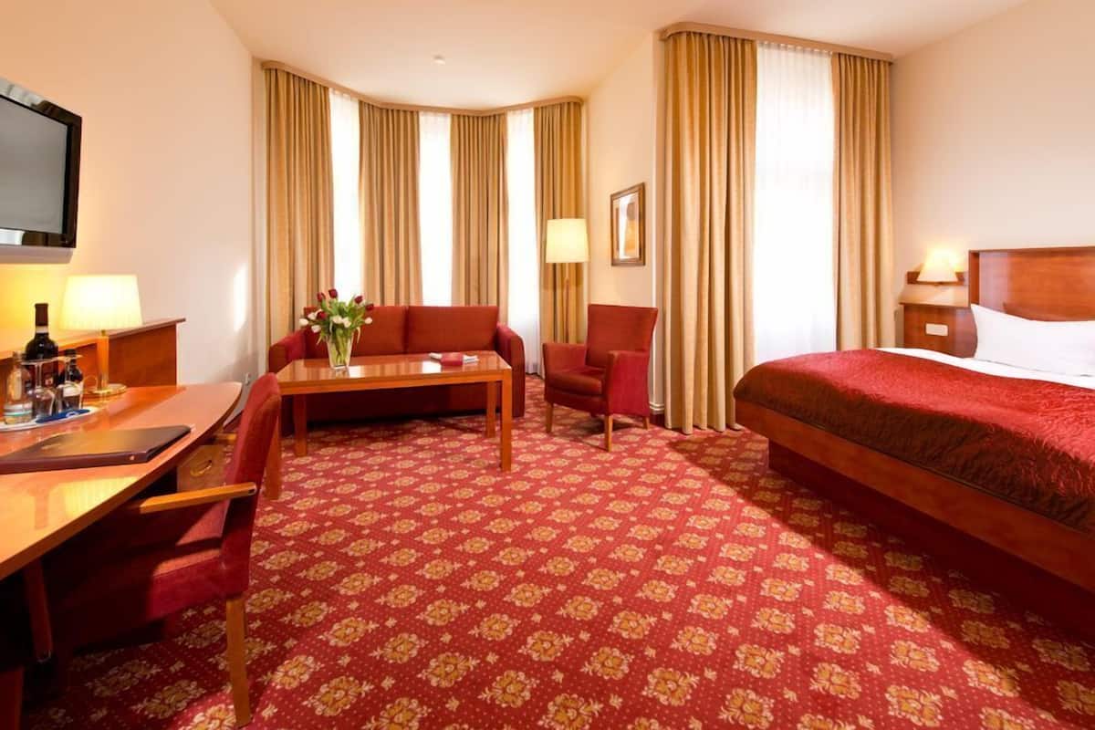 A hotel room with a bed, a desk with a chair, a small table with a vase of flowers, and a television on the wall.