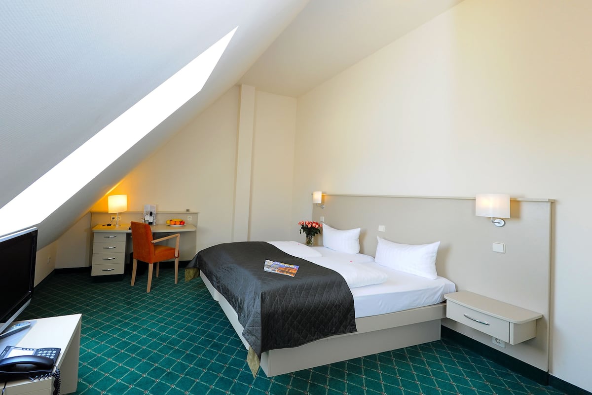 A hotel room with a bed, desk, chair, and a small table with a lamp.