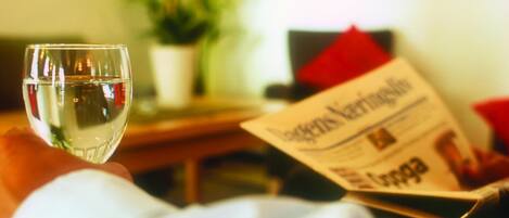A person holding a glass of water, reading a newspaper, with a plant and a red pillow in the background.