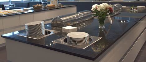Montaje de bufé con platos apilados, dispensador de bebidas y un florero sobre una encimera pulida.
