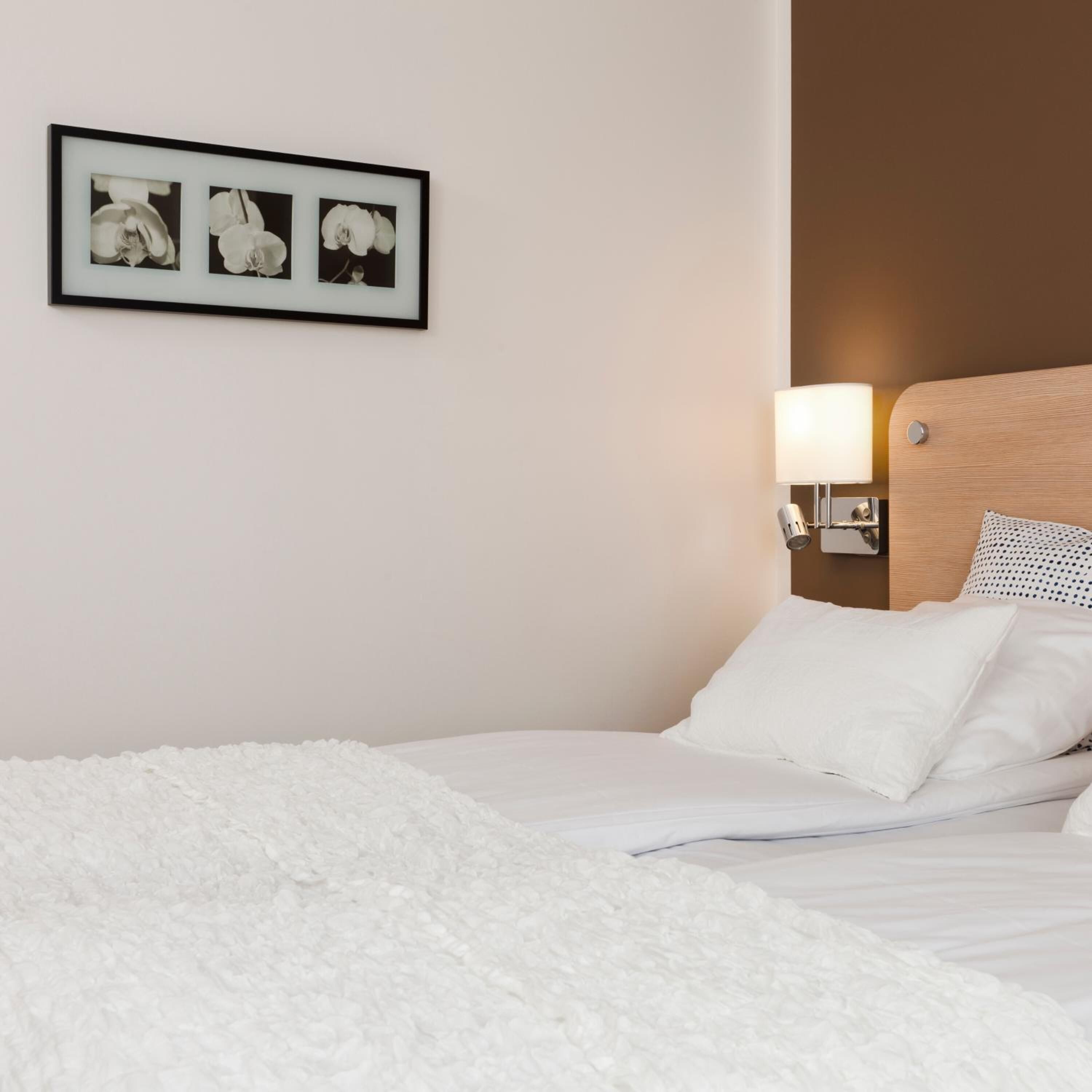 A neatly made bed with white bedding, a wall-mounted picture of flowers, and a bedside lamp.