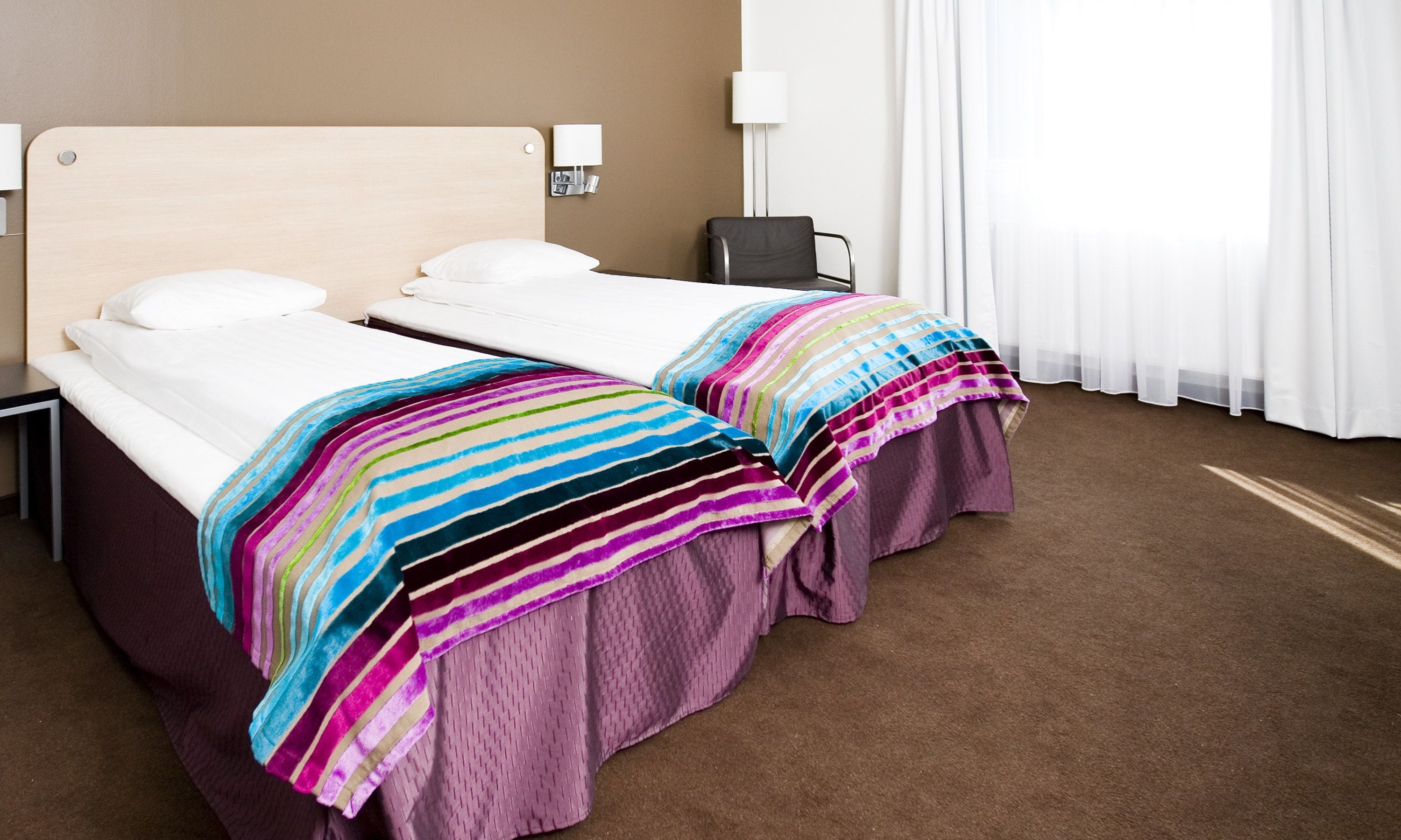 Habitación de hotel con dos camas, cada una con una manta a rayas, una mesita de noche, una lámpara y una silla.