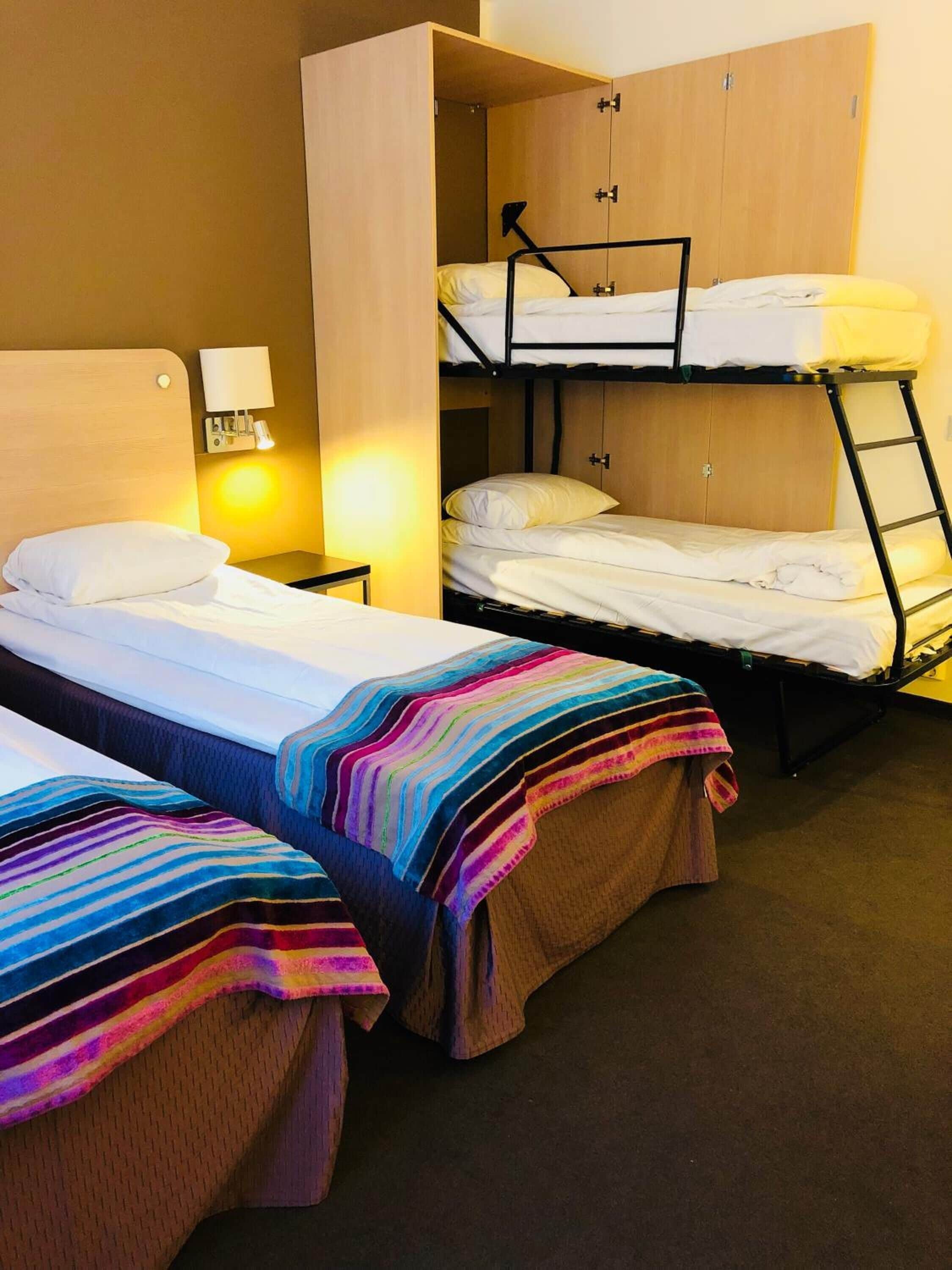 Une chambre de dortoir avec des lits superposés, une armoire en bois, une table de chevet avec une lampe et une couverture rayée colorée sur l’un des lits.