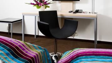 A modern office chair with a black backrest and metallic legs, a wooden desk with a telephone and a small plant, and a colorful striped sofa in the foreground.