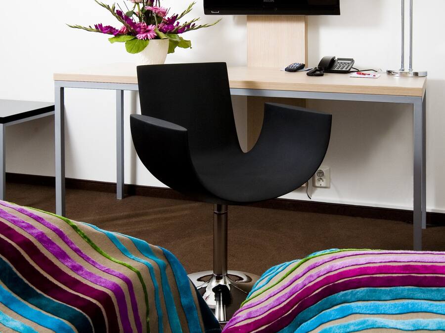 A modern office chair with a black backrest and metallic legs, a wooden desk with a telephone and a small plant, and a colorful striped sofa in the foreground.