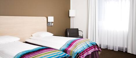 Habitación de hotel con dos camas individuales, un escritorio, una silla, una lámpara y una ventana con cortinas traslúcidas.