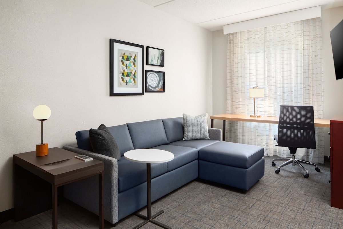 Ein modernes Hotelzimmer mit einem blauen Ecksofa, einem kleinen runden Tisch, einem Schreibtisch mit Stuhl und gerahmten Kunstwerken an der Wand.