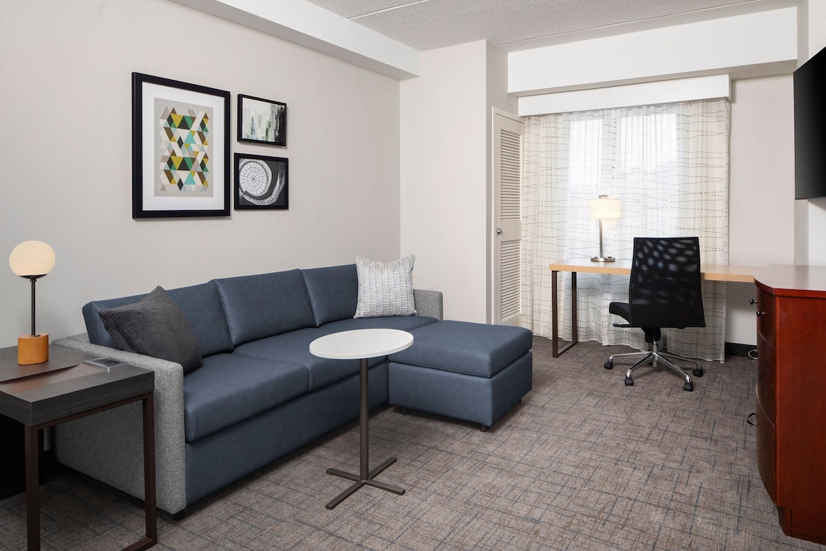 Ein modernes Hotelzimmer mit einer Eckcouch, einem kleinen runden Tisch, einem Schreibtisch mit Stuhl und Bildern an den Wänden.