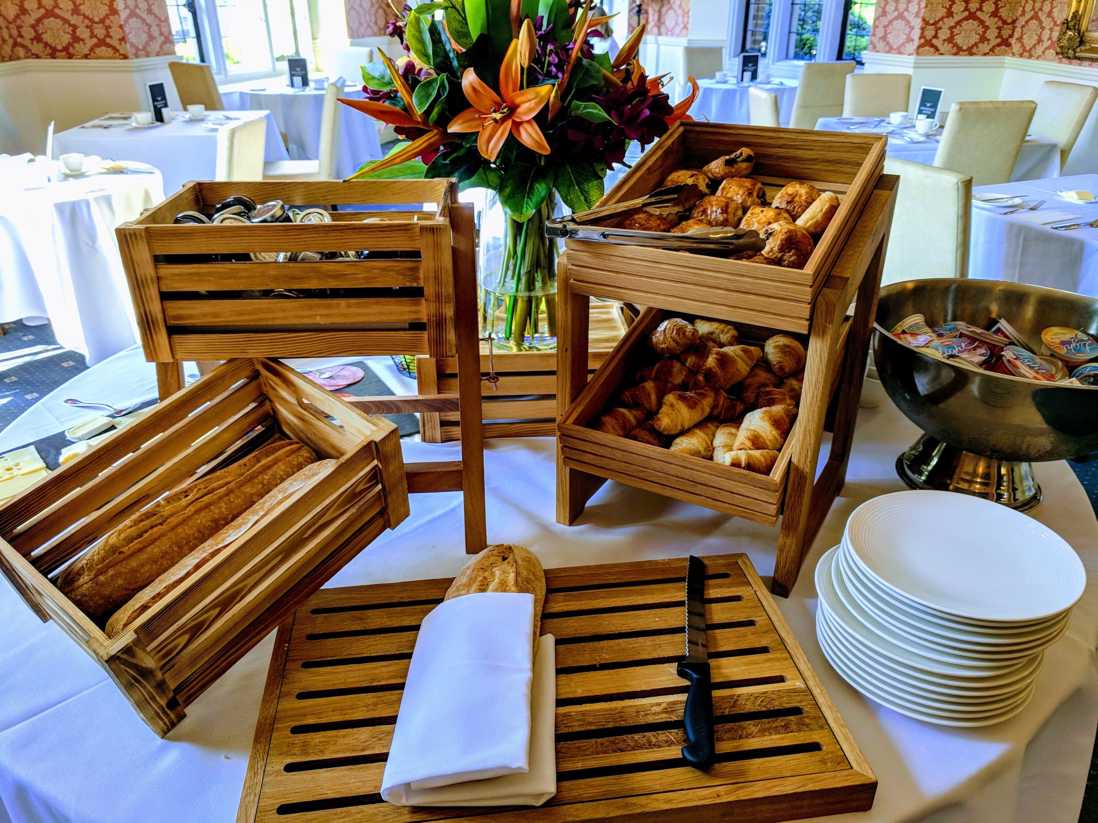 A dining setup with wooden crates containing pastries and bread, a knife on a wooden board, and a bowl of utensils.