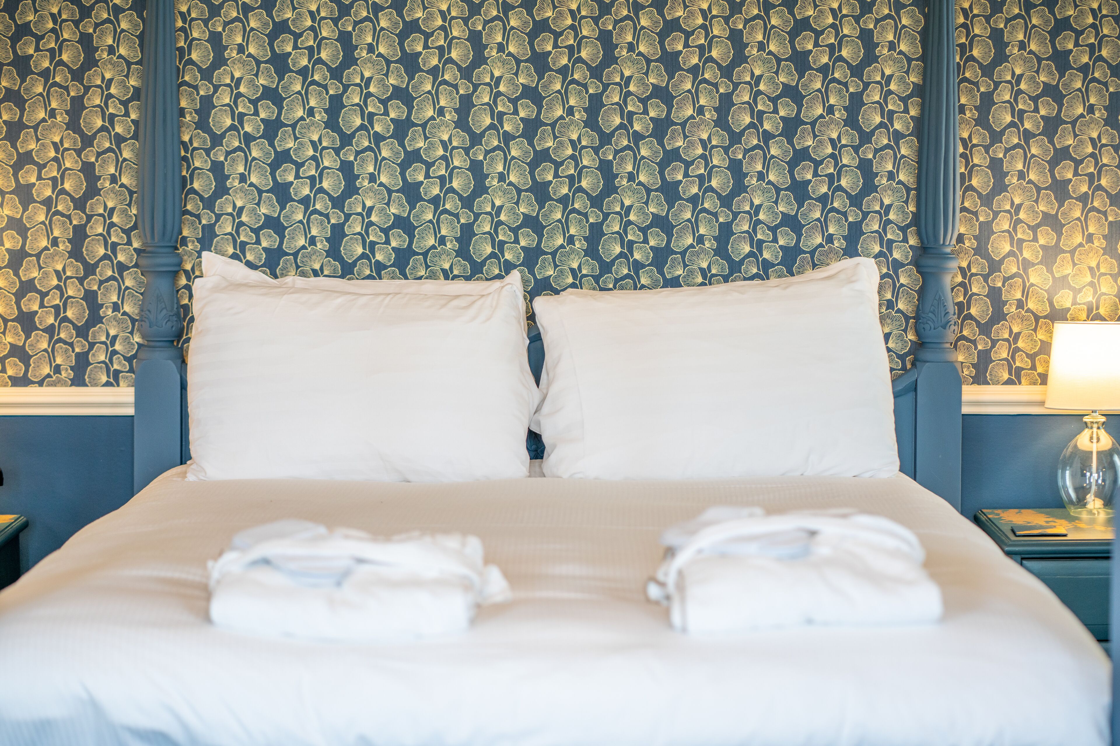 A neatly made bed with white linens and towels, a blue headboard, and a patterned wallpaper.