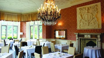 A dining room with multiple tables set for service, a large chandelier, and a fireplace with a decorative mantle.