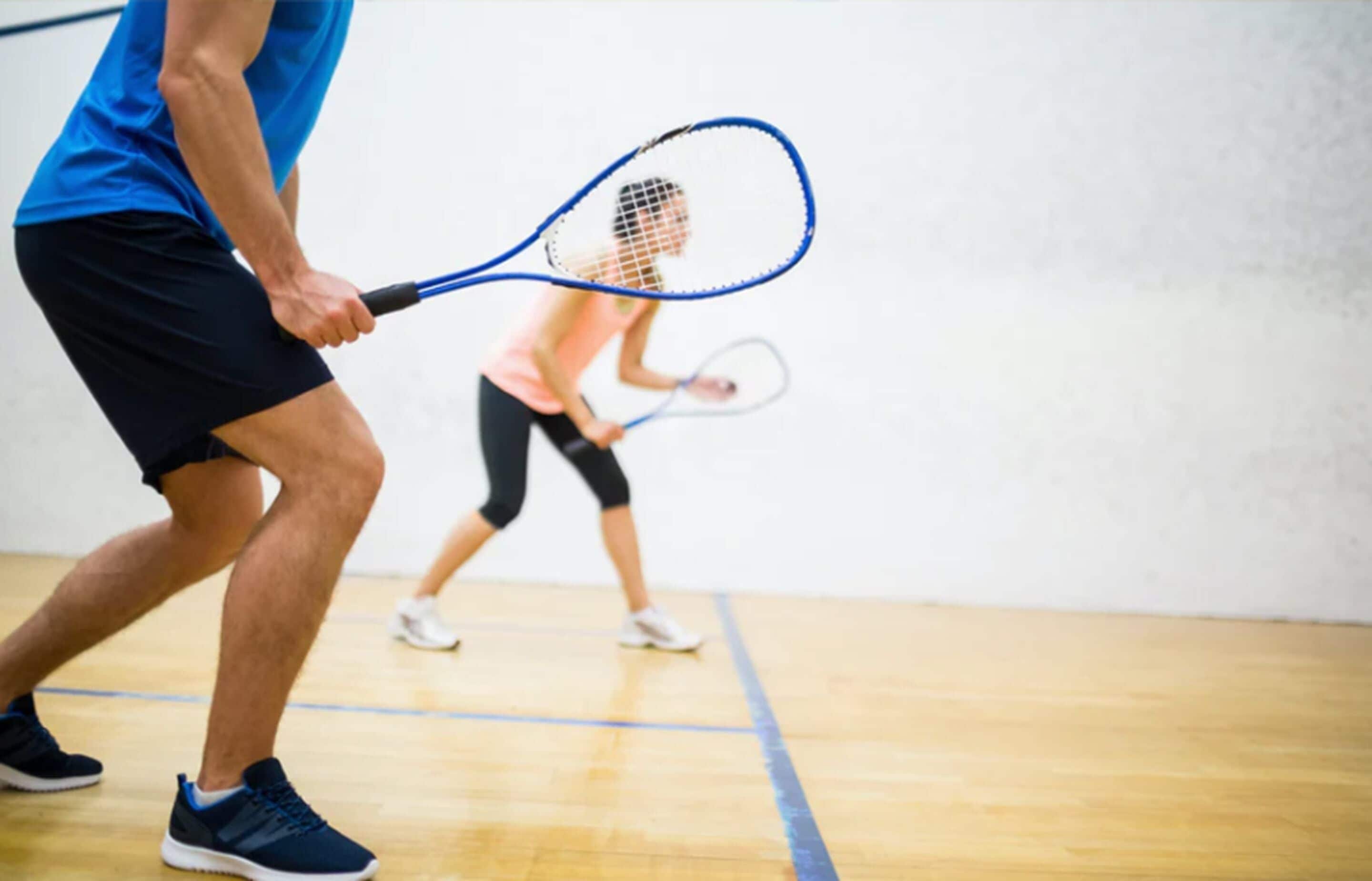 A person holding a tennis racket in a gymnasium with a wooden floor and a white wall.